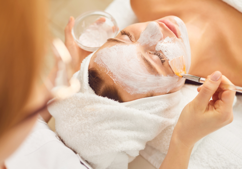 A person receiving a facial treatment, with a mask applied by an esthetician using a brush. The person has a towel wrapped around their head and eyes closed.