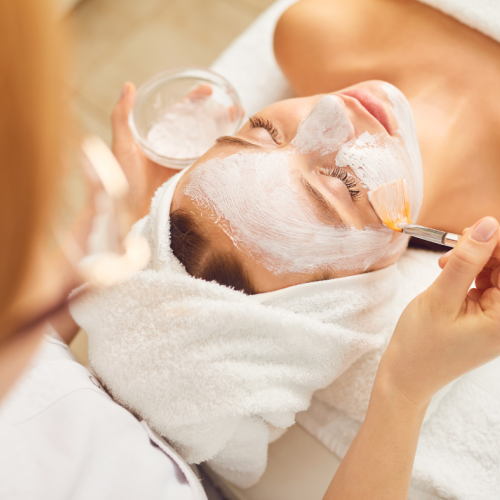 A person receiving a facial treatment, with a mask applied by an esthetician using a brush. The person has a towel wrapped around their head and eyes closed.