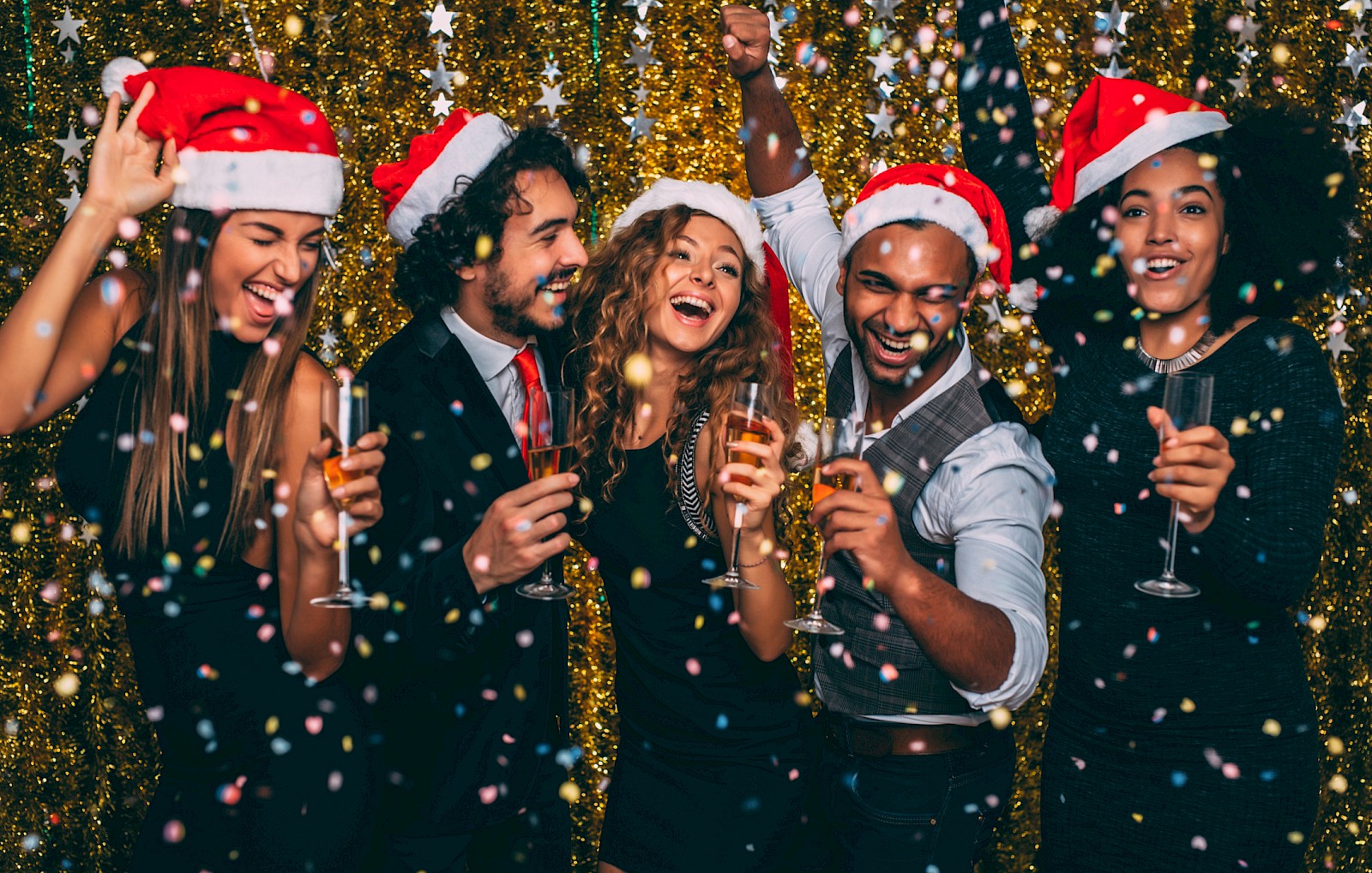 A festive group wearing Santa hats, celebrating with drinks and confetti in front of a glittery backdrop.
