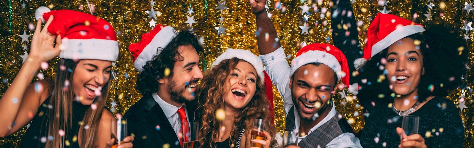 A festive group wearing Santa hats, celebrating with drinks and confetti in front of a glittery backdrop.