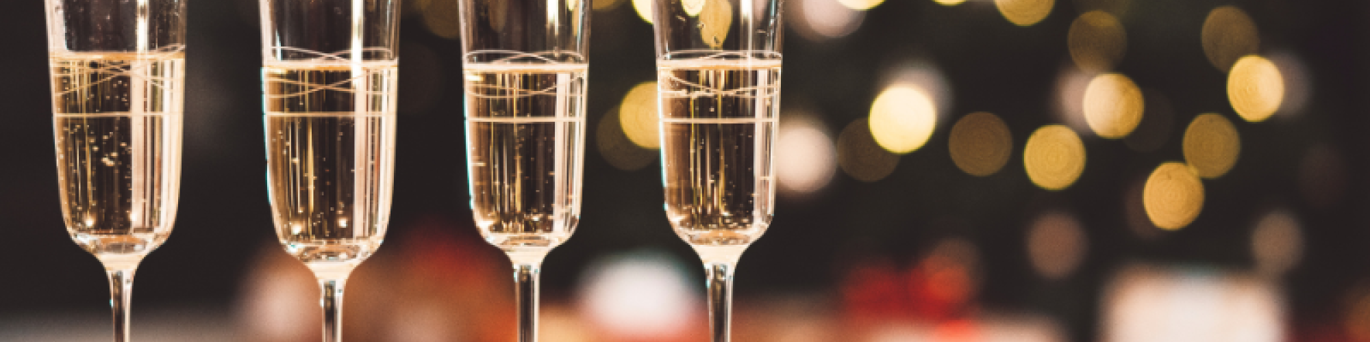 Four filled champagne flutes are on a wooden table with a blurred Christmas tree and presents in the background, creating a festive atmosphere.