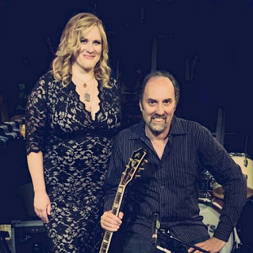 A woman in a black lace dress and a man holding a guitar at a music event.