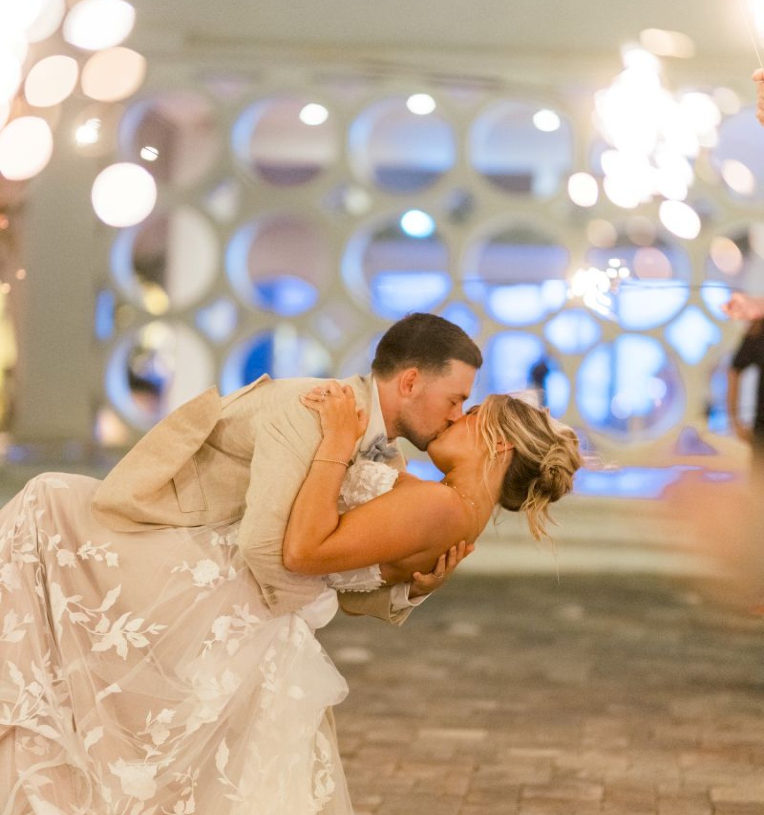 A newlywed couple shares a romantic kiss during their wedding celebration with sparklers in a festive outdoor setting.