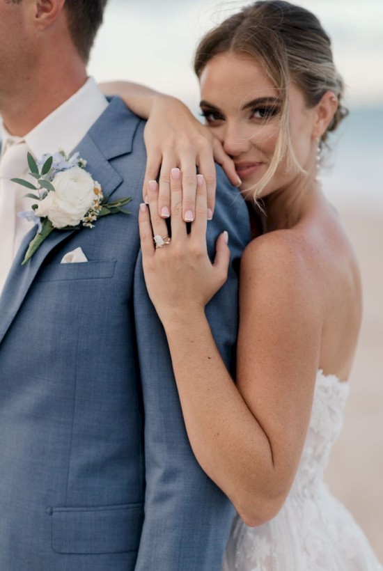 A bride and groom embrace, showing off her engagement ring, with the bride smiling behind the groom, dressed in wedding attire at an outdoor setting.