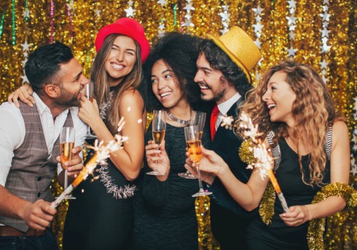 A group of five friends celebrate with sparklers and champagne, smiling and toasting at a festive party with a gold sequin backdrop.