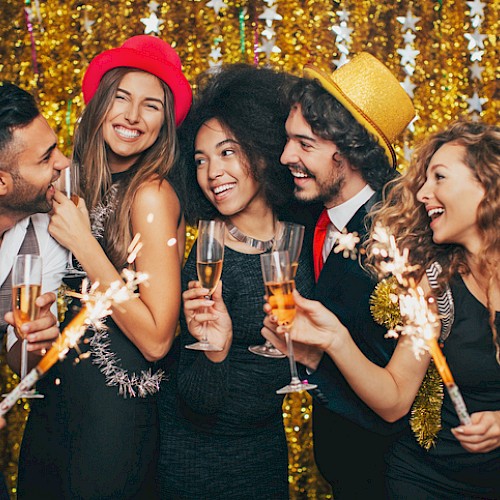 A group of five friends celebrate with sparklers and champagne, smiling and toasting at a festive party with a gold sequin backdrop.