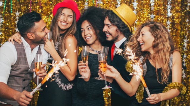 A group of five friends celebrate with sparklers and champagne, smiling and toasting at a festive party with a gold sequin backdrop.