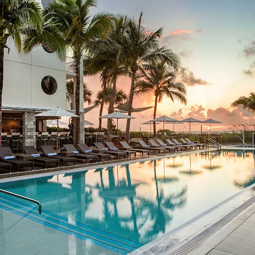 A resort-style swimming pool with lounge chairs, palm trees, and a sunset sky reflecting on calm water, perfect for sunset cocktails.