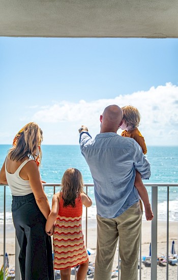 A family stands on a balcony, overlooking a sunny beach and ocean. The sky is mostly clear with a few clouds in view.