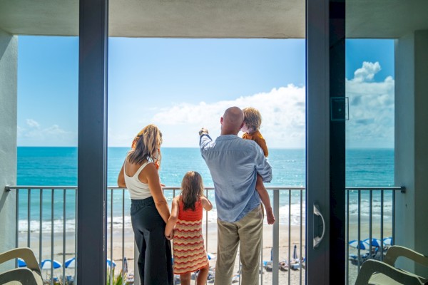 A family of four stands on a balcony, overlooking a sunny beach and ocean view, with one child pointing towards the horizon.