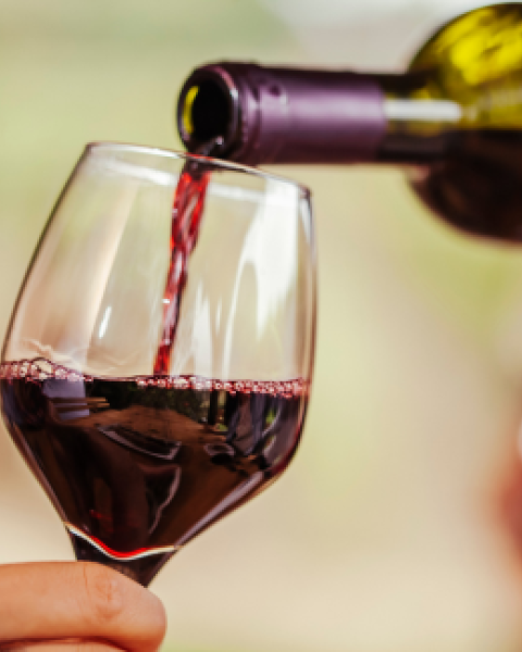 A hand holds a wine glass while red wine is being poured from a bottle, filling the glass.