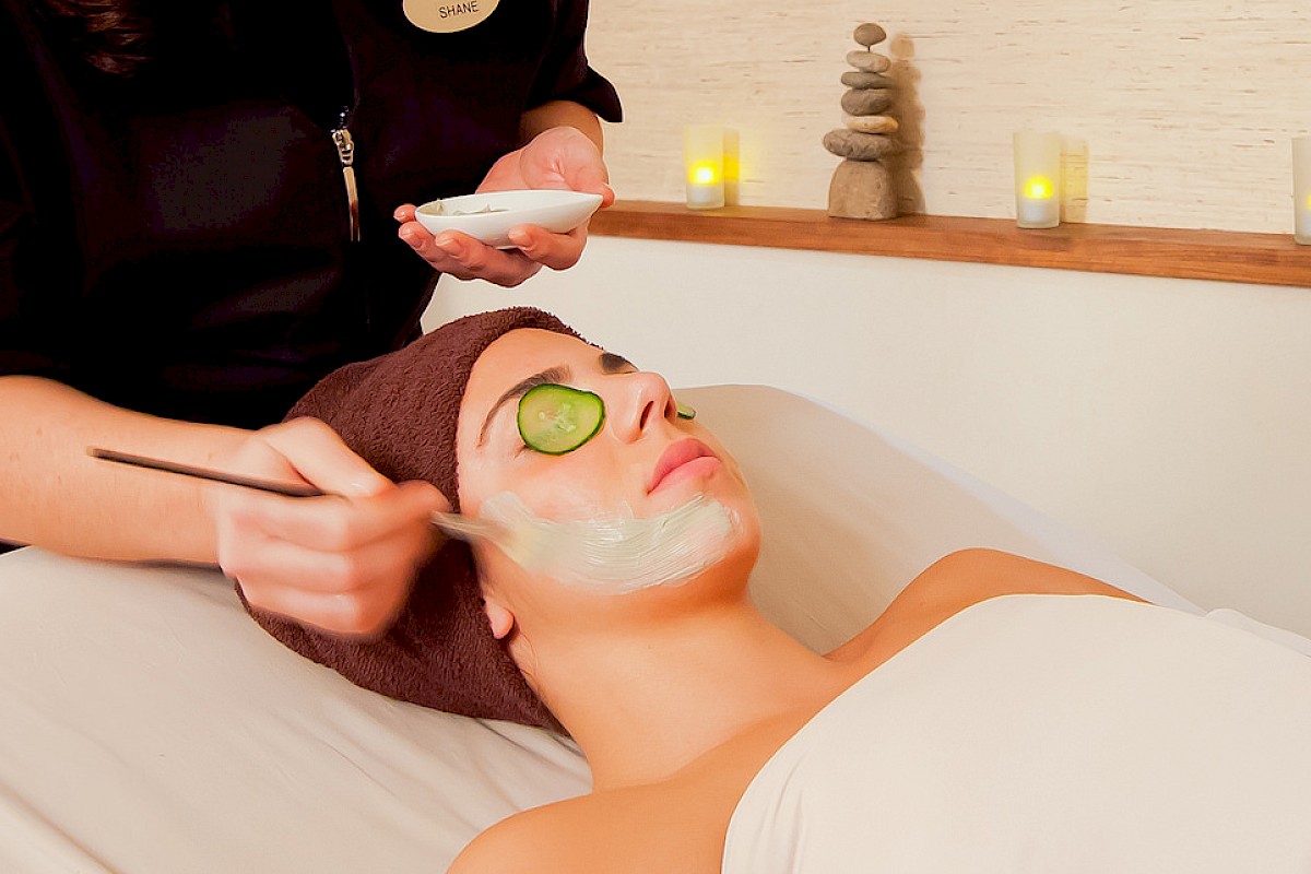 A woman is getting a facial treatment with cucumber slices on her eyes at a spa, while a practitioner applies a mask to her face.