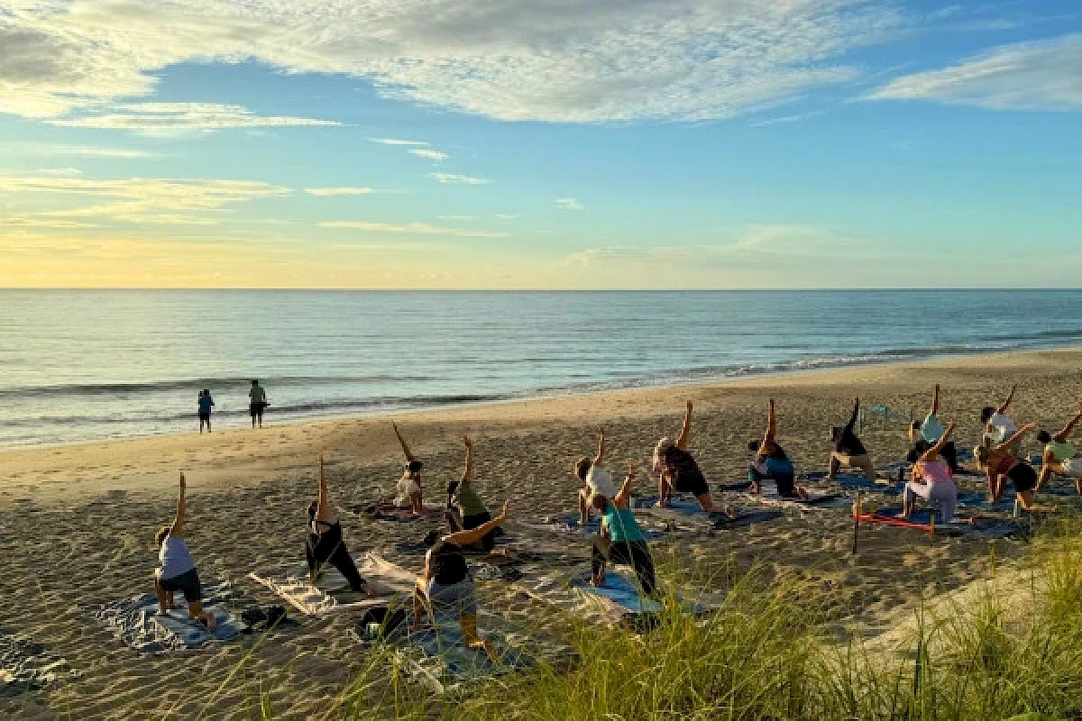 People are practicing yoga on the beach at sunrise or sunset near the ocean, with a few walking along the shoreline.