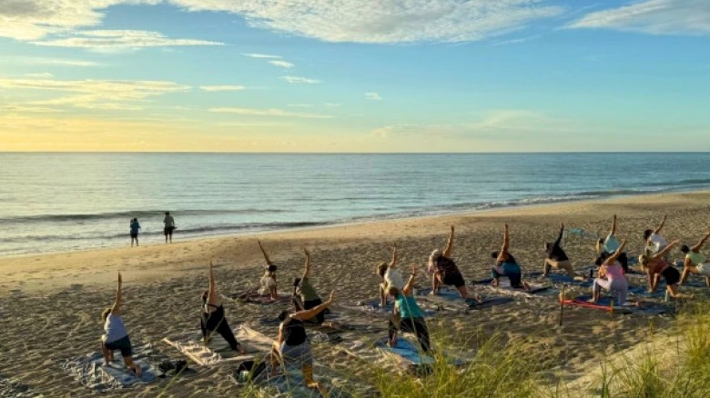 People are practicing yoga on the beach at sunrise or sunset near the ocean, with a few walking along the shoreline.