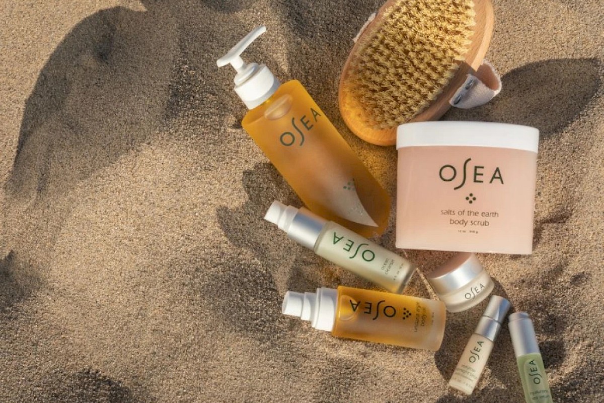 Beach skincare and body products, including body scrub, oils, and a brush, are arranged on the sand for a relaxing beach day.