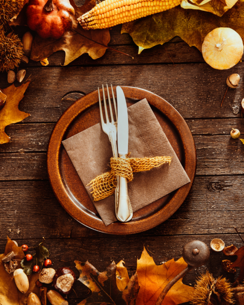 A rustic autumn table setting with a plate, utensils, corn, pumpkins, leaves, and nuts, creating a warm, seasonal atmosphere.