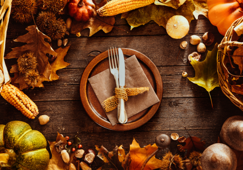 A rustic autumn table setting with a plate, utensils, corn, pumpkins, leaves, and nuts, creating a warm, seasonal atmosphere.