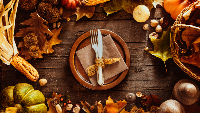 A rustic autumn table setting with a plate, utensils, corn, pumpkins, leaves, and nuts, creating a warm, seasonal atmosphere.