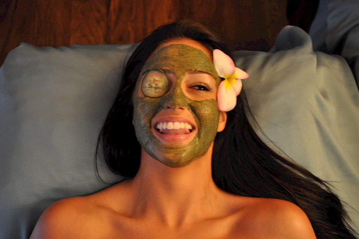 A woman with a green facial mask and an eye patch, lying on a pillow with a flower in her hair, smiling and sticking out her tongue.