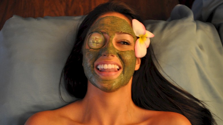 A woman with a green facial mask and an eye patch, lying on a pillow with a flower in her hair, smiling and sticking out her tongue.