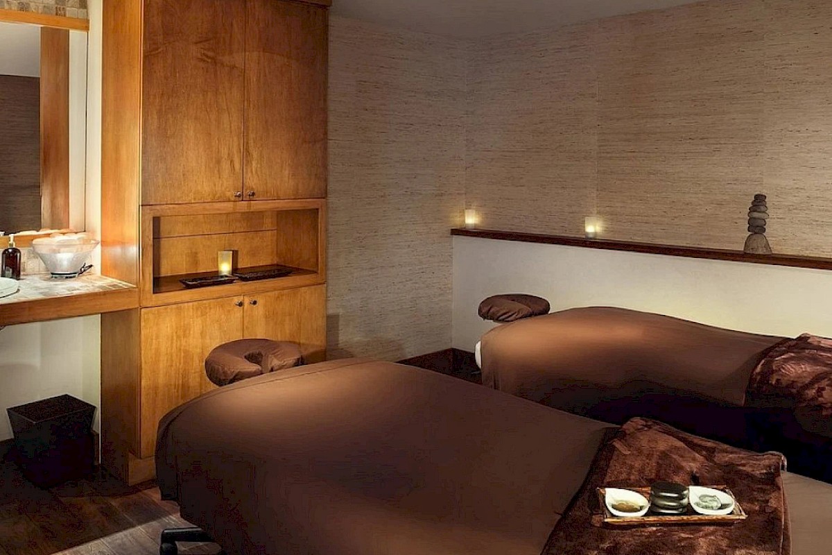 The image shows a cozy bedroom with two beds, wooden furniture, warm lighting, and a minimalist, peaceful ambiance.
