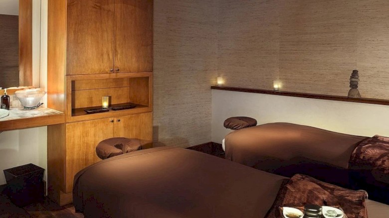 The image shows a cozy bedroom with two beds, wooden furniture, warm lighting, and a minimalist, peaceful ambiance.