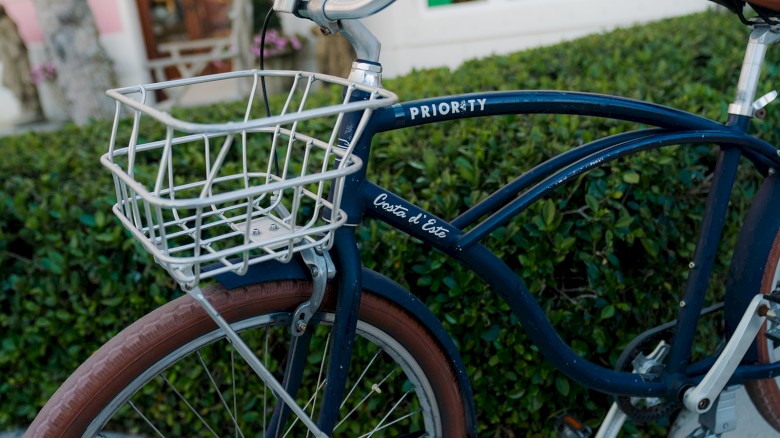 A blue bicycle with a white wire basket parked in front of green bushes and a building with large windows.