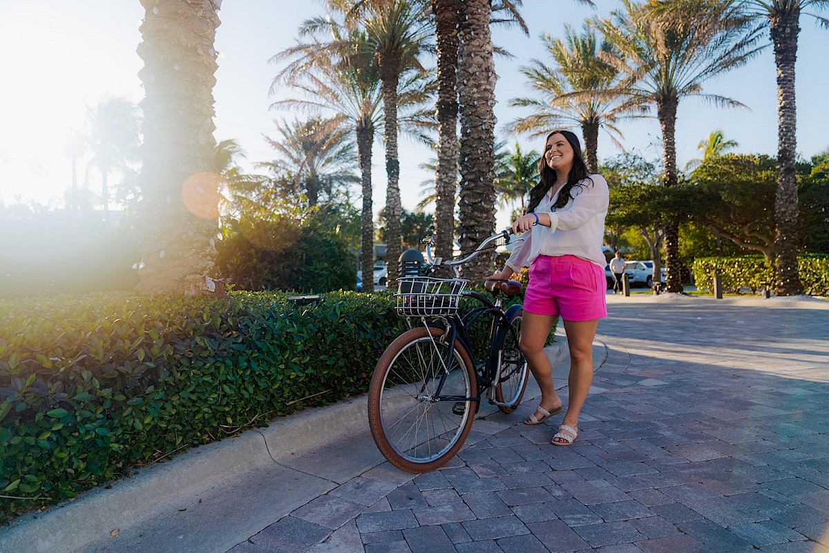 A woman in pink shorts and a white top stands with her bike near palm trees on a sunny day.