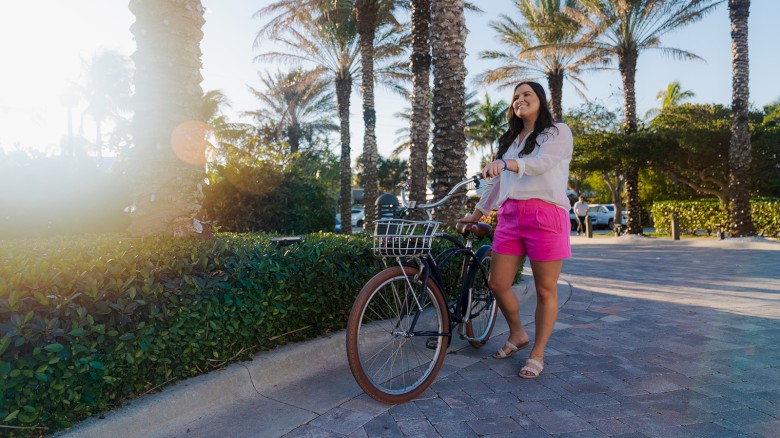 A woman in pink shorts and a white top stands with her bike near palm trees on a sunny day.