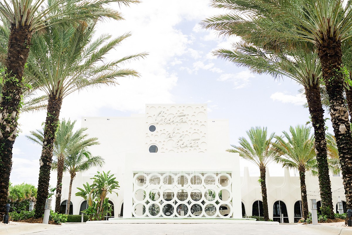 A white building with circular patterns, surrounded by tall palm trees and a partly cloudy sky in the background.