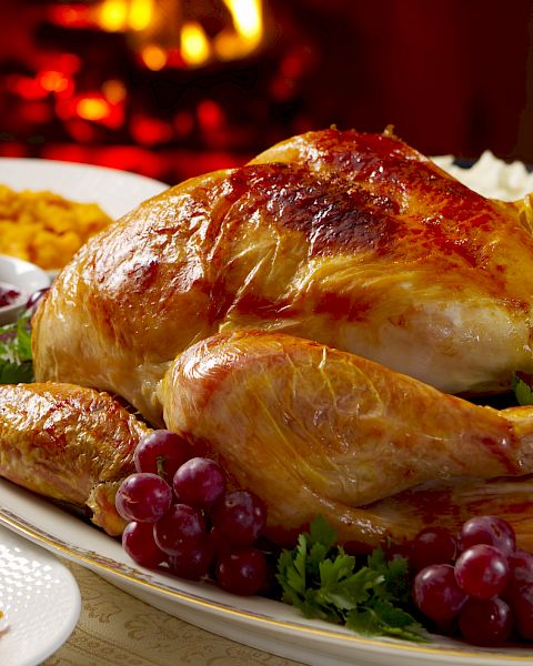 The image shows a roasted turkey garnished with grapes and parsley, accompanied by sides like mashed sweet potatoes and cranberry sauce near a fireplace.