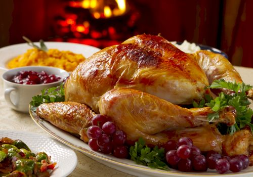 The image shows a roasted turkey garnished with grapes and parsley, accompanied by sides like mashed sweet potatoes and cranberry sauce near a fireplace.