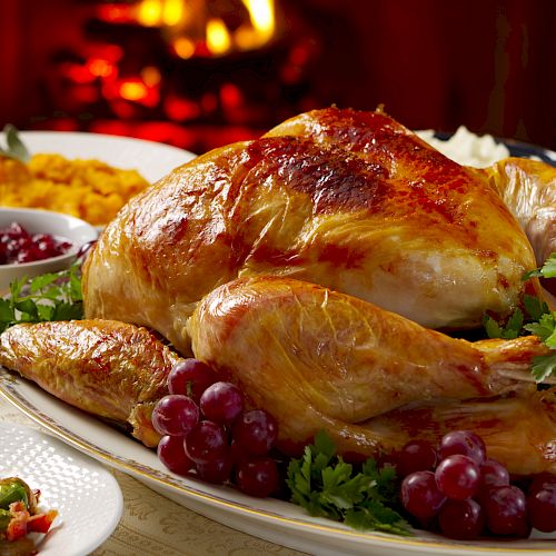 The image shows a roasted turkey garnished with grapes and parsley, accompanied by sides like mashed sweet potatoes and cranberry sauce near a fireplace.