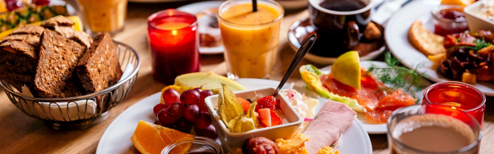 An elaborate breakfast spread with eggs, fruit, bread, and beverages on a wooden table, featuring juices and candles for a cozy setting.