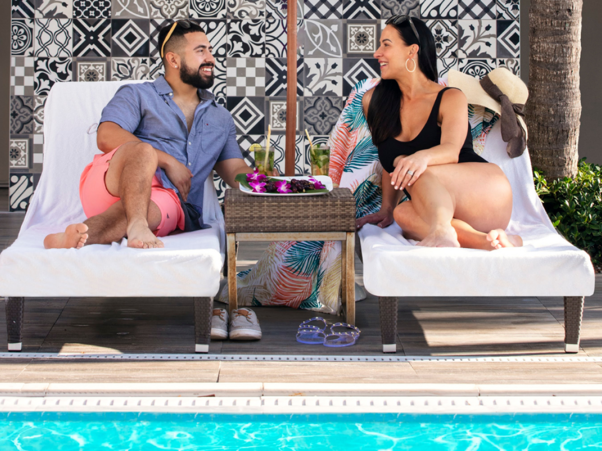 A man and woman relax on poolside lounge chairs, chatting and smiling, with a table and drinks between them.