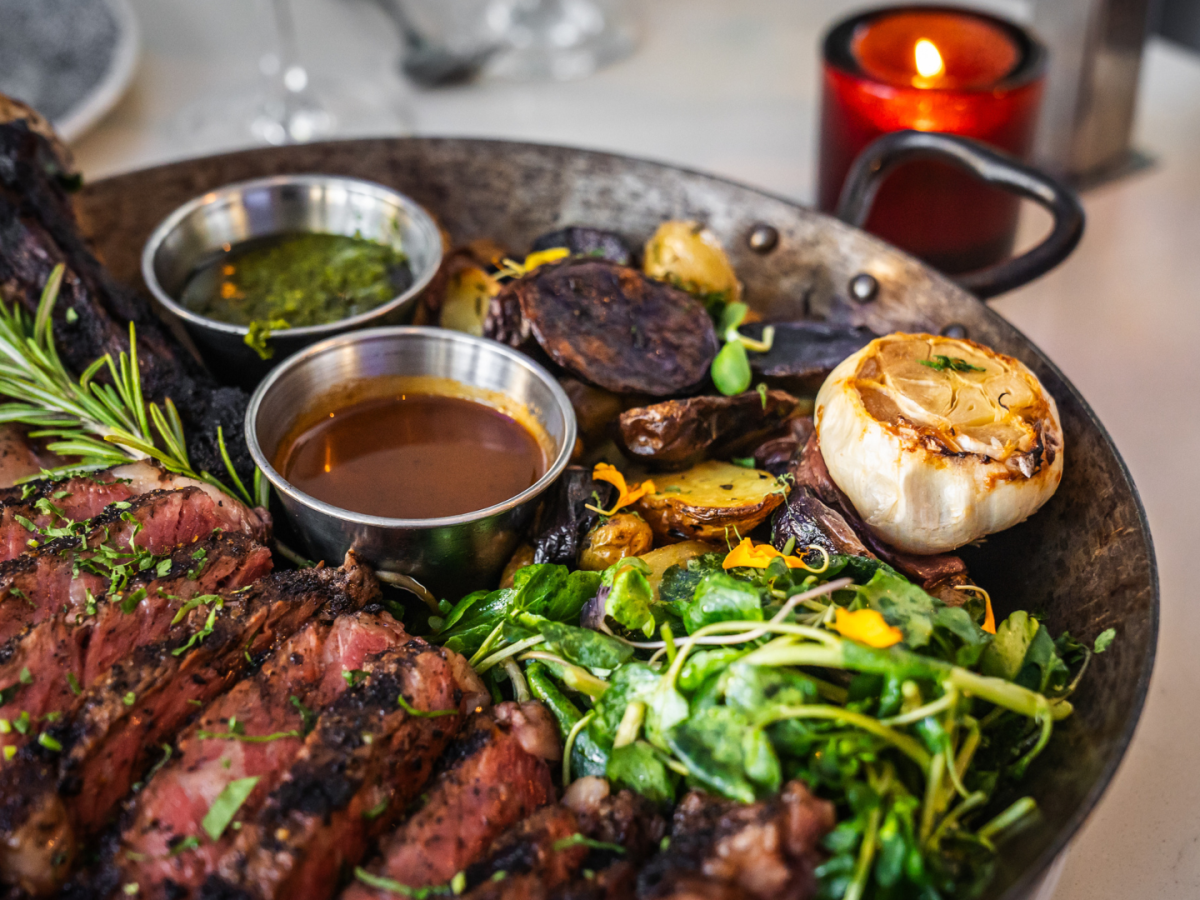 A platter with grilled steak, roasted garlic, potatoes, greens, and two sauces garnished with rosemary, alongside a lit red candle on the table.