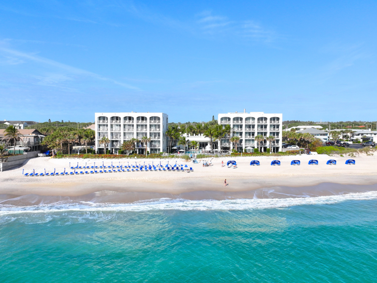 A beachside setting with two modern buildings, lined with blue umbrellas and loungers on the sandy shore, facing a calm sea.