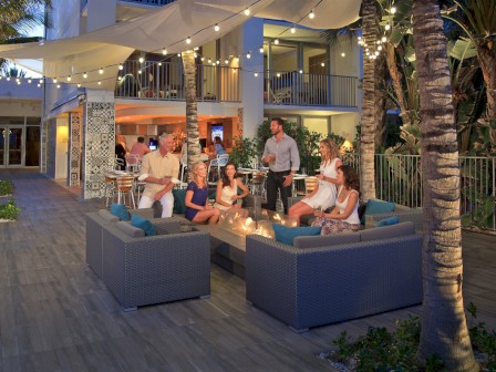 People are relaxing and socializing around a fire pit at an outdoor lounge with palm trees, string lights, and a modern building in the background.