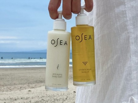 The image shows a hand holding two bottles of Ocean skincare products on a beach, with the ocean and sky in the background.
