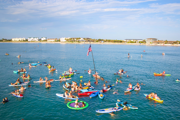 Many people are enjoying a day on the water with paddleboards, kayaks, and floaties near a coastal city.