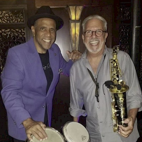 Two musicians posing together: one in a purple jacket with conga drums, the other in a light shirt holding a saxophone, smiling at the camera.