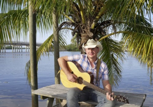 A man in a cowboy hat sits on a wooden dock by the water, playing an acoustic guitar with palm trees in the background.