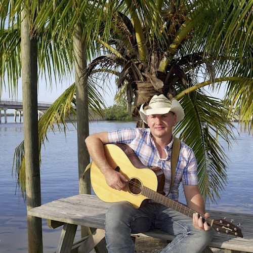 A man in a cowboy hat sits on a wooden dock by the water, playing an acoustic guitar with palm trees in the background.