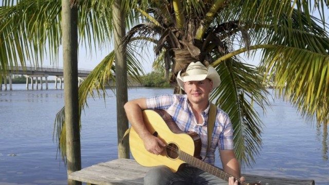 A man in a cowboy hat sits on a wooden dock by the water, playing an acoustic guitar with palm trees in the background.