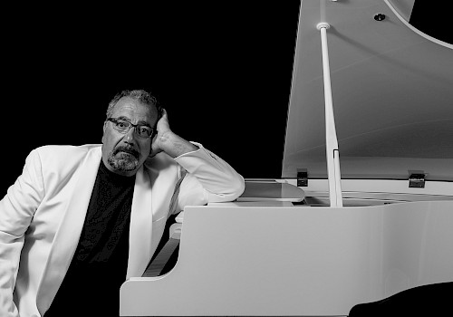 A man in a white suit leans on a white grand piano against a black background, gazing thoughtfully.