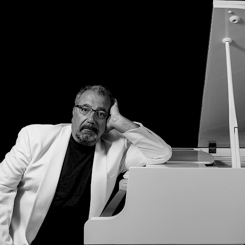 A man in a white suit leans on a white grand piano against a black background, gazing thoughtfully.