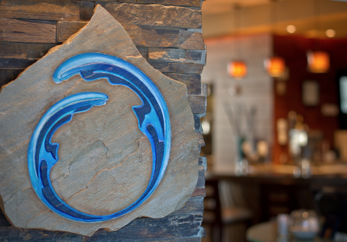 This image shows a stylized blue logo on a stone wall with a blurred restaurant interior in the background.