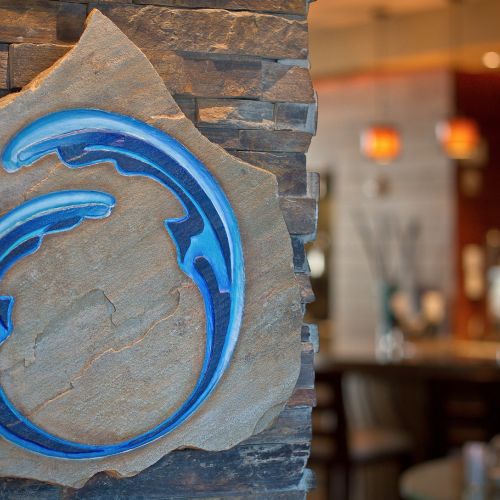 This image shows a stylized blue logo on a stone wall with a blurred restaurant interior in the background.