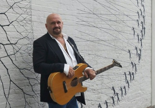 A person with a guitar stands against a mural-covered wall depicting small figures and lines, dressed in a dark jacket and jeans.