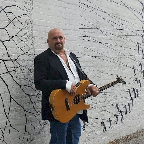 A person with a guitar stands against a mural-covered wall depicting small figures and lines, dressed in a dark jacket and jeans.